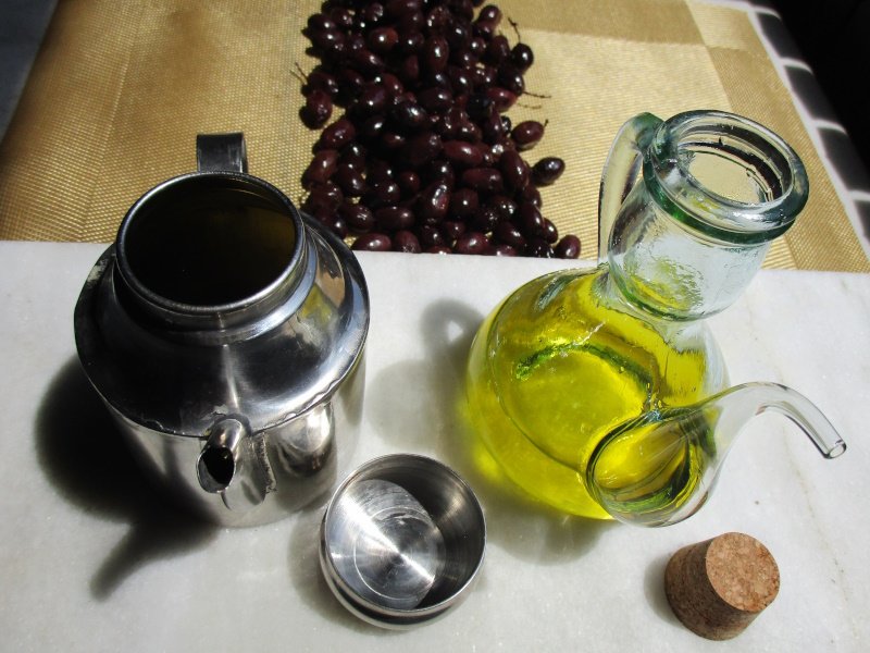 ACEITE DE OLIVA EN LAS OLLAS FRANCISCANAS II Dos aceiteras de cocinas caseras españolas del siglo XXI, una de vidrio reciclado, la segunda de acero inoxidable. El diseño remonta a la época de al-Andalus. En la mesa hay aceitunas Empeltre adobadas. © Vicky Hayward, 2025
